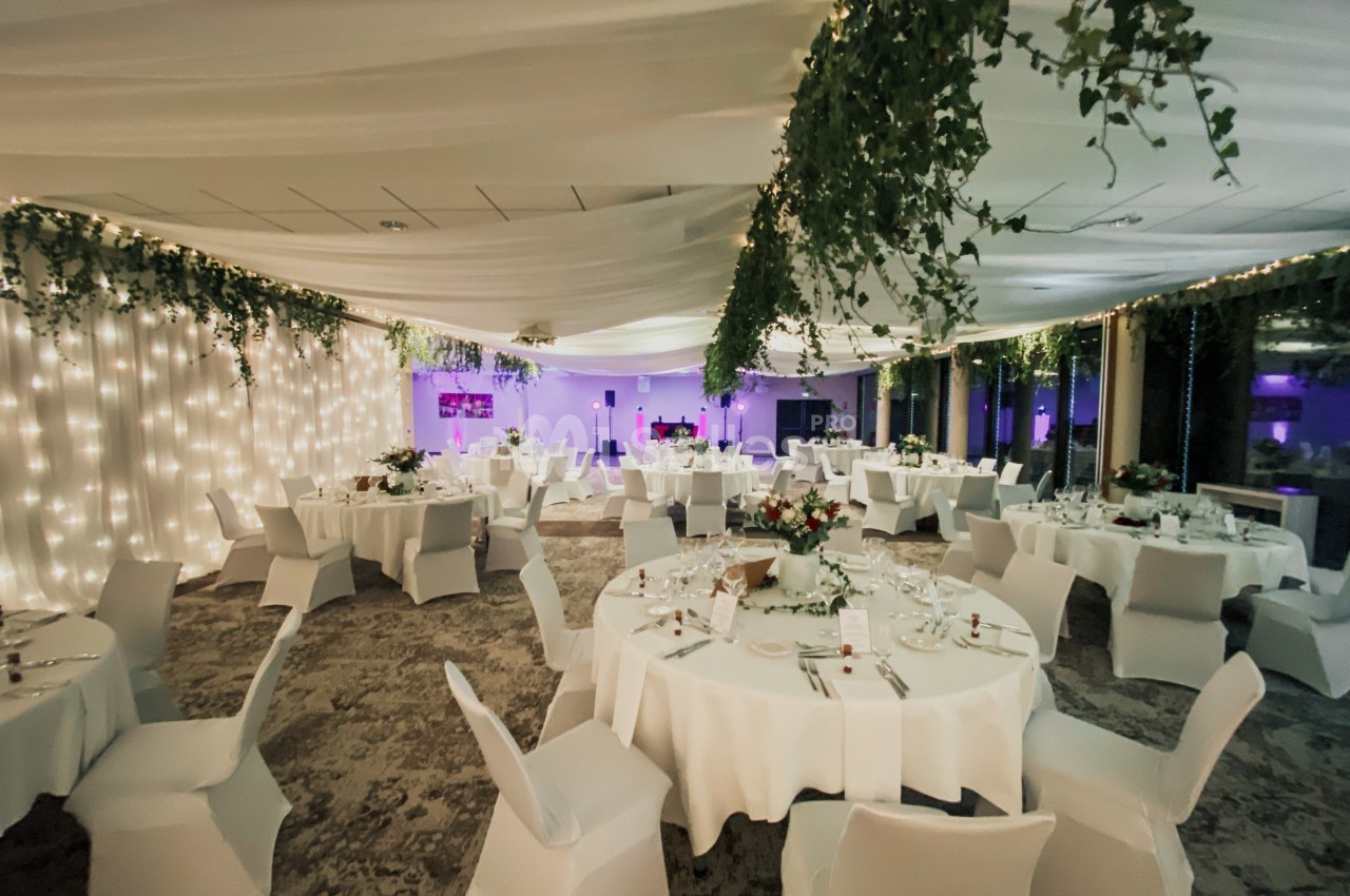 Salle de réception décorée avec des guirlandes lumineuses, des nappes blanches et des arrangements floraux sur les tables.