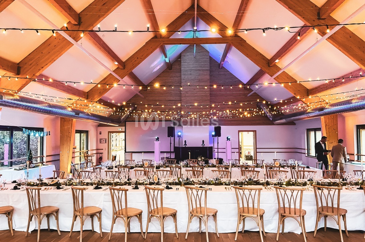 Grande salle décorée pour un événement, avec tables dressées, chaises en bois et guirlandes lumineuses suspendues.