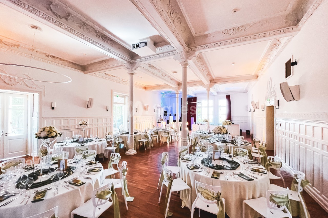 Salle de réception élégante avec tables rondes dressées, décorations florales et lumière naturelle entrant par les fenêtres.