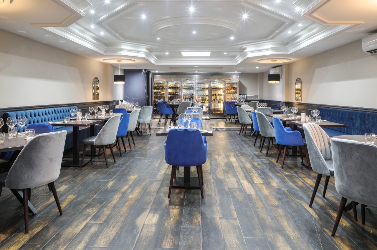 Salle de restaurant élégante avec tables dressées, chaises en velours bleu et gris, et éclairage moderne.