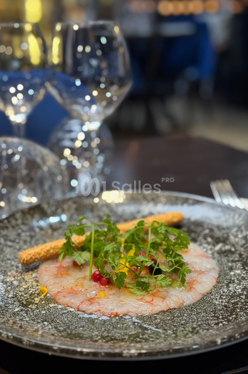 Carpaccio de crevettes décoré de fines herbes, baies rouges et accompagné d'un gressin, servi sur une assiette noire.