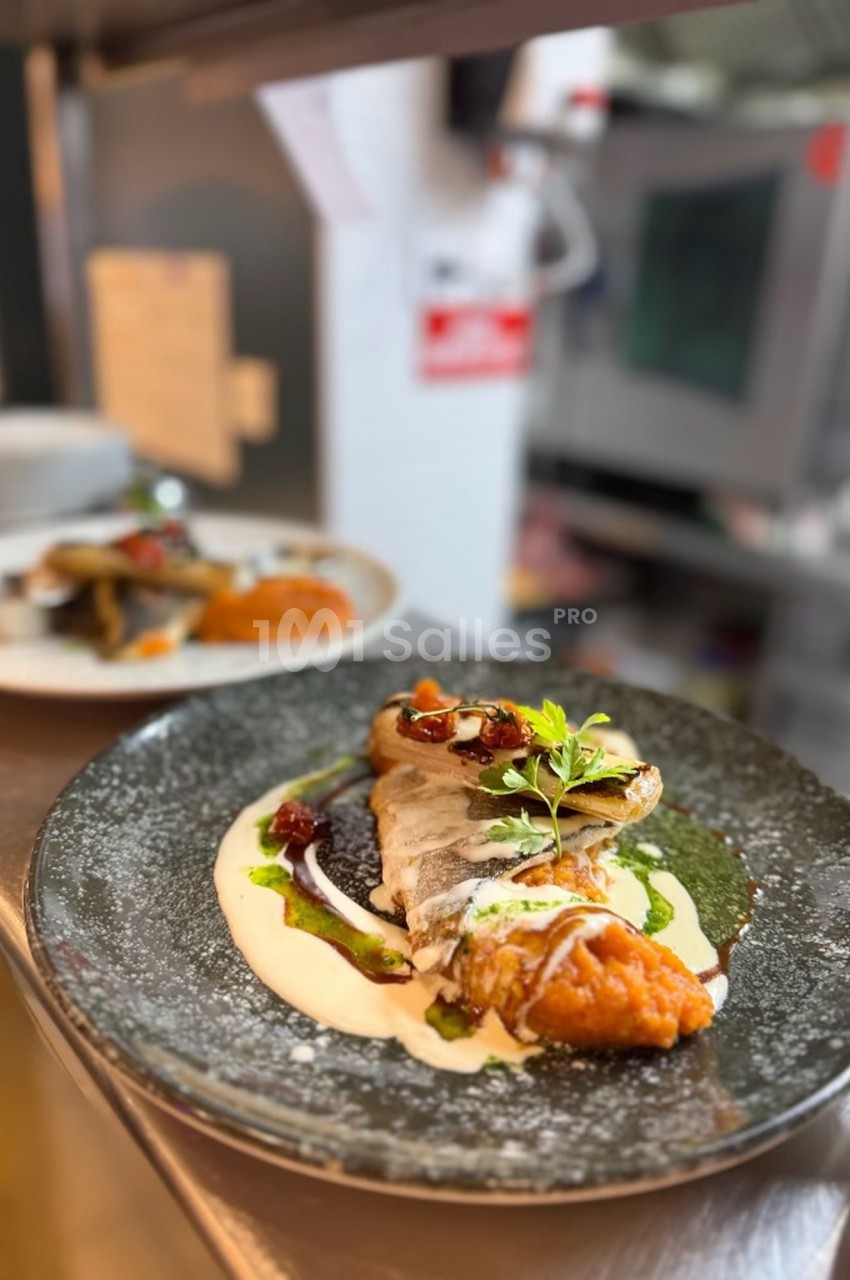 Assiette de poisson grillé avec purée, sauce crémeuse, légumes et herbes fraîches, servie dans une cuisine professionnelle.