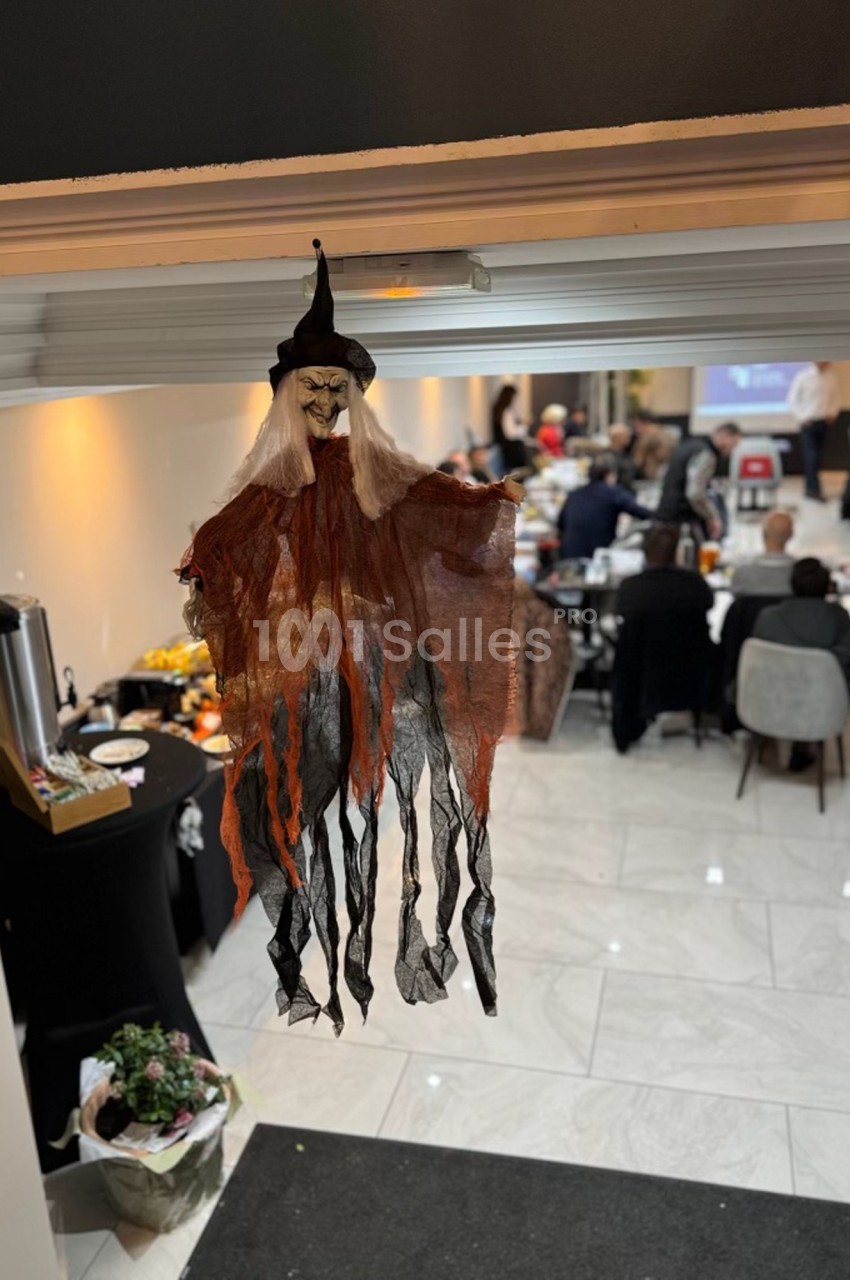 Décoration de sorcière suspendue dans une salle lumineuse où des personnes sont rassemblées autour de tables.