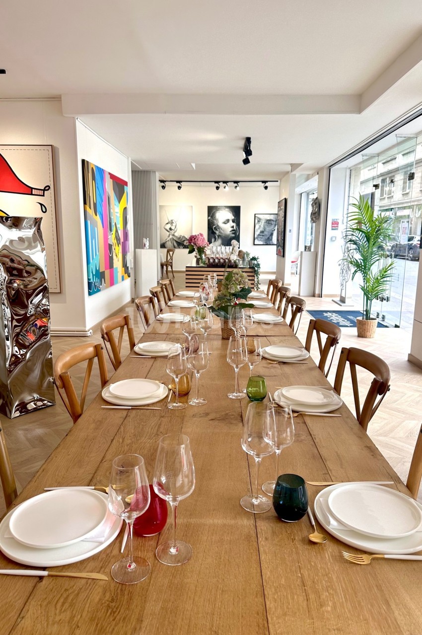 Grande table en bois dressée avec vaisselle et verres, entourée de chaises, dans une salle lumineuse décorée d'œuvres d'art.