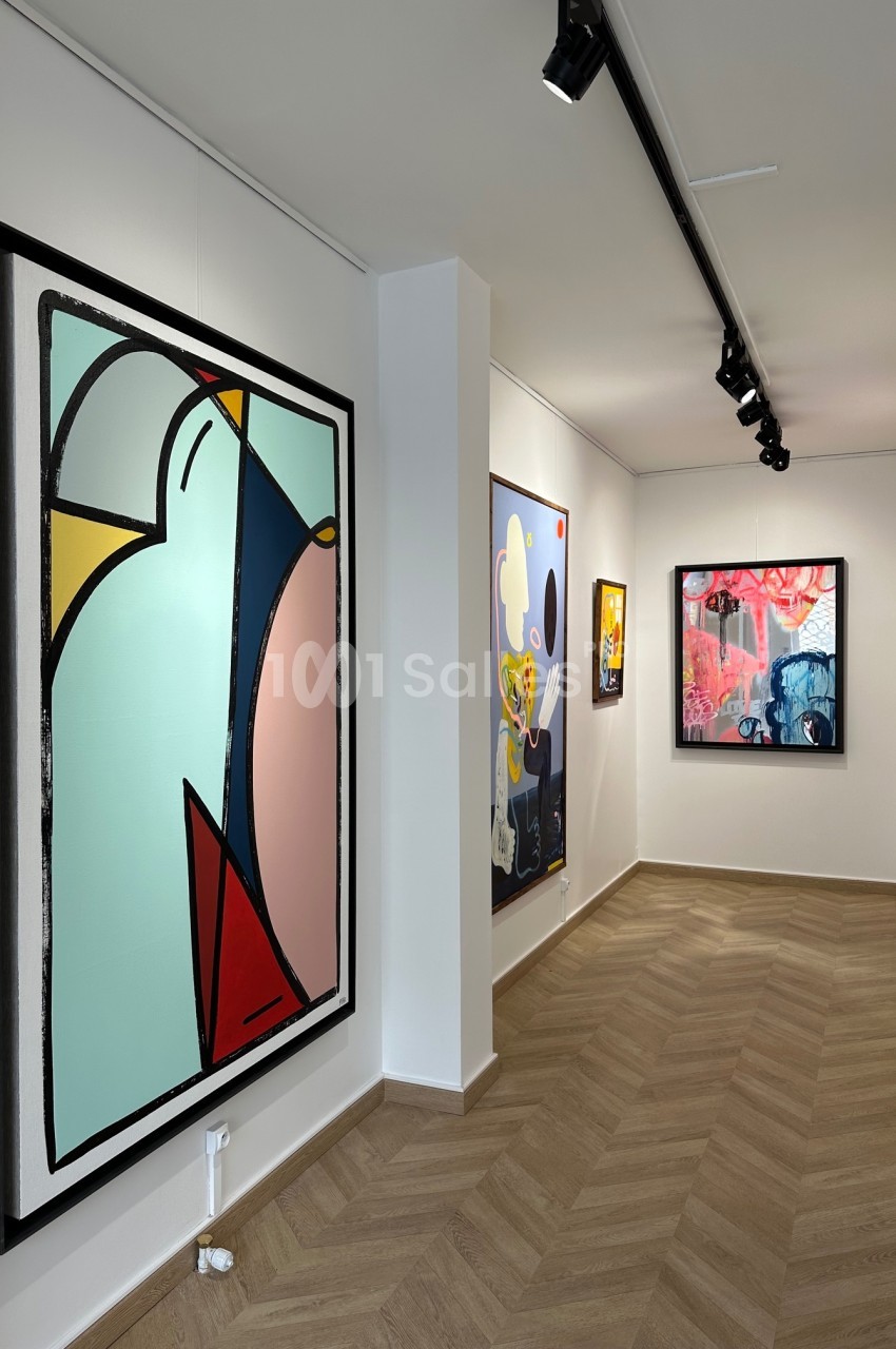 Vue d'une galerie d'art avec des tableaux modernes et colorés accrochés sur des murs blancs, éclairés par des spots.