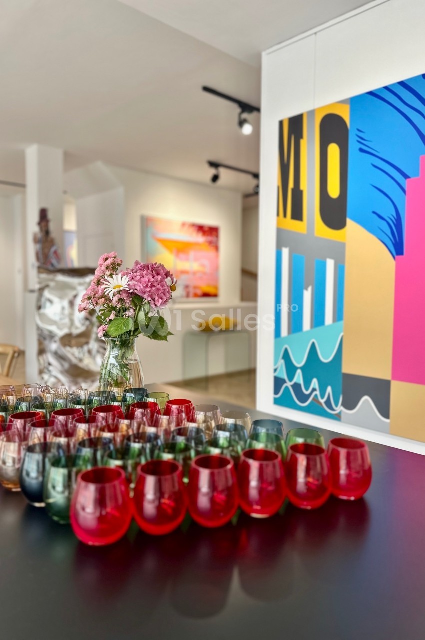 Vase de fleurs roses sur une table noire entourée de verres colorés, dans une galerie d'art moderne lumineuse.