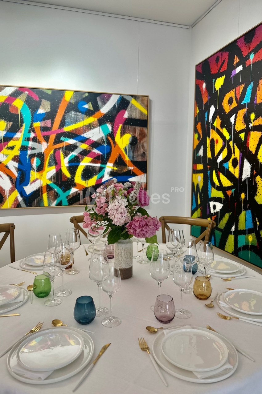 Table dressée avec vaisselle colorée et bouquet de fleurs, entourée de chaises, devant des tableaux abstraits colorés.