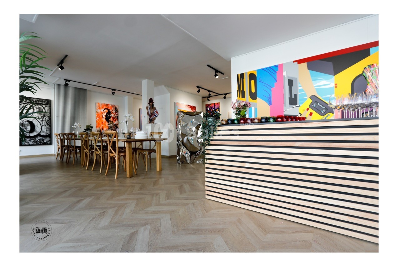 Salle lumineuse avec parquet en chevrons, table en bois, œuvres d'art modernes aux murs et comptoir à l'avant-plan.