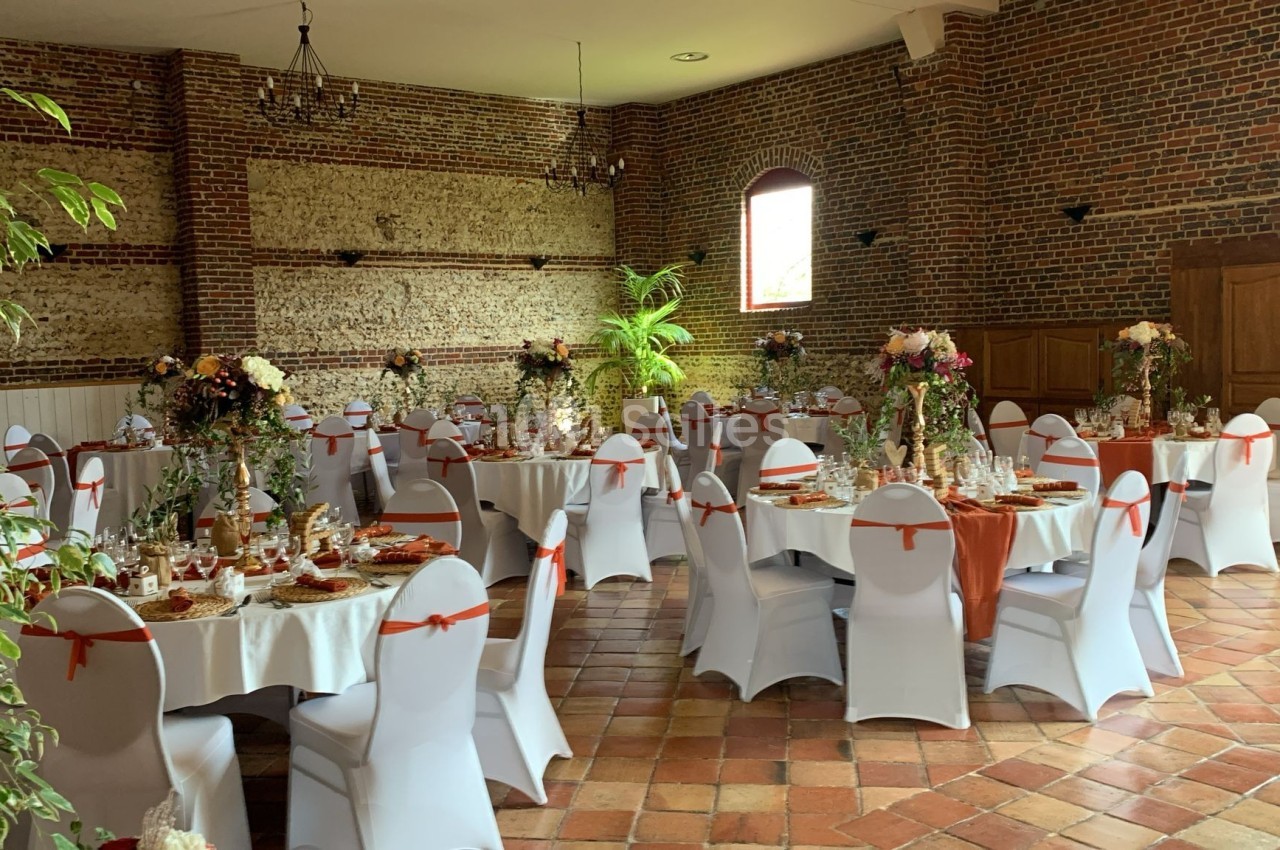Salle de réception décorée avec des tables rondes, nappes blanches, chaises ornées de nœuds orange et compositions florales.