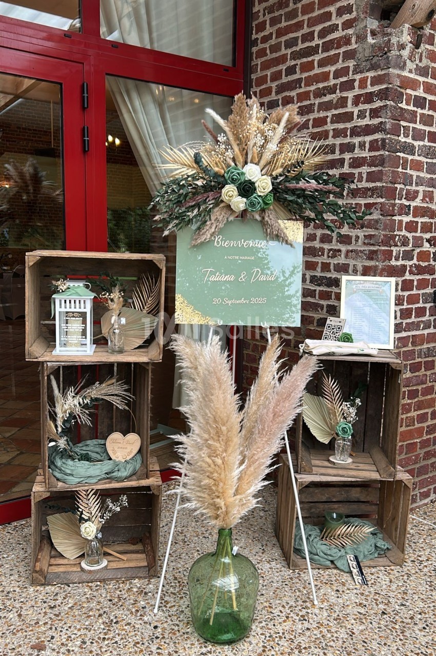 Décoration de mariage avec caisses en bois, fleurs séchées, vase en verre et panneau de bienvenue devant une porte rouge.