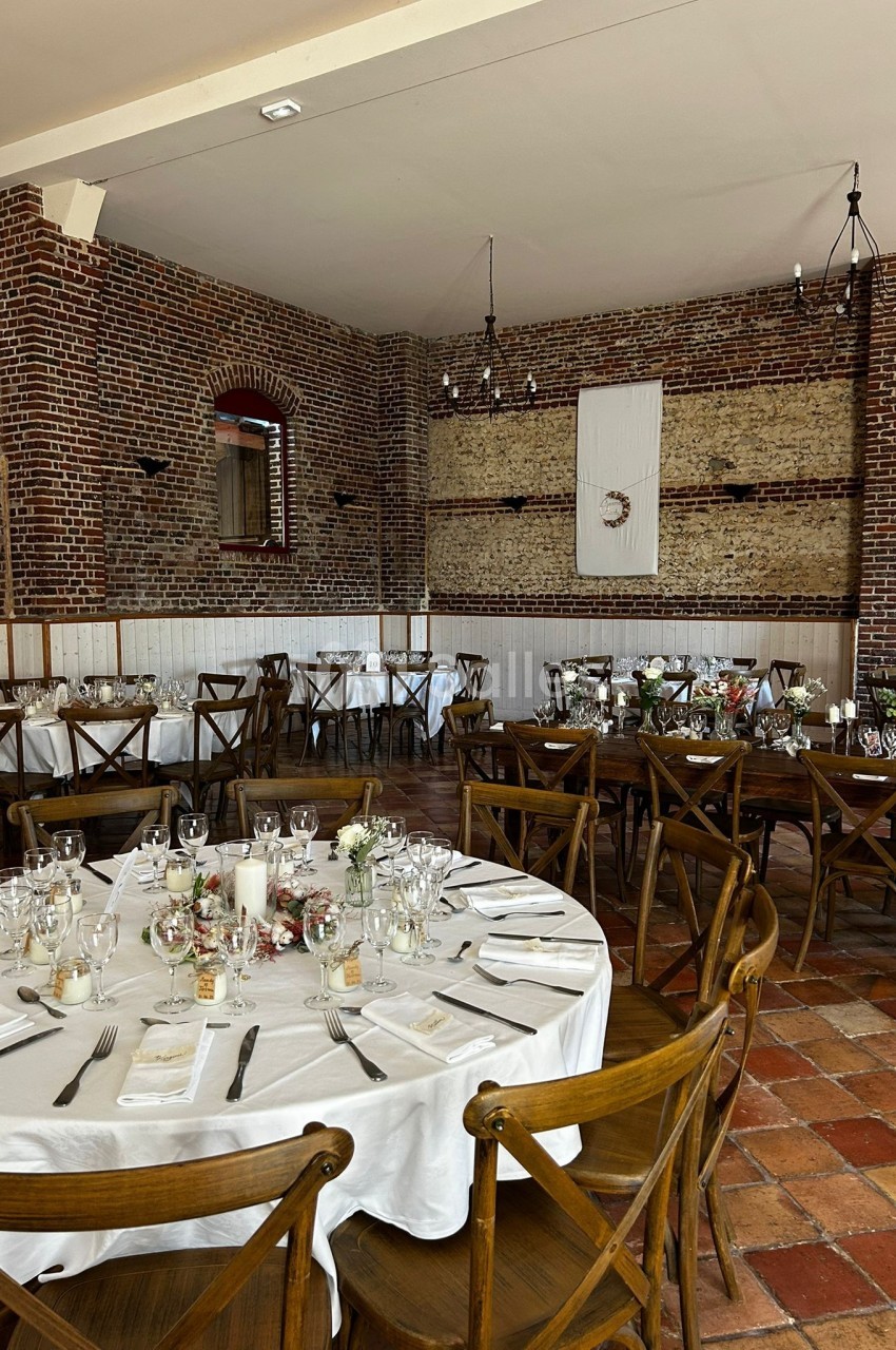 Salle de réception avec murs en briques, tables rondes dressées avec nappes blanches et chaises en bois.