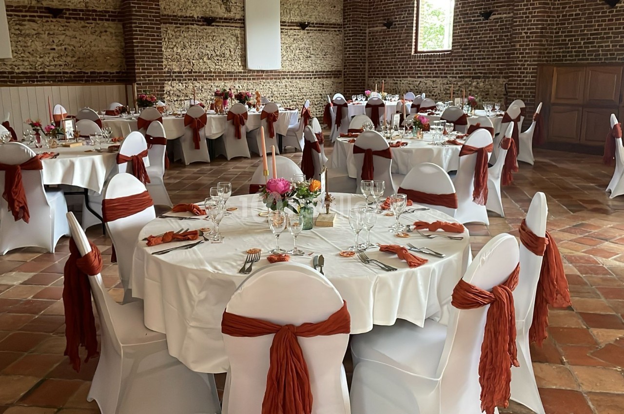 Salle de réception décorée avec des tables rondes, nappes blanches et chaises ornées de nœuds rouges.
