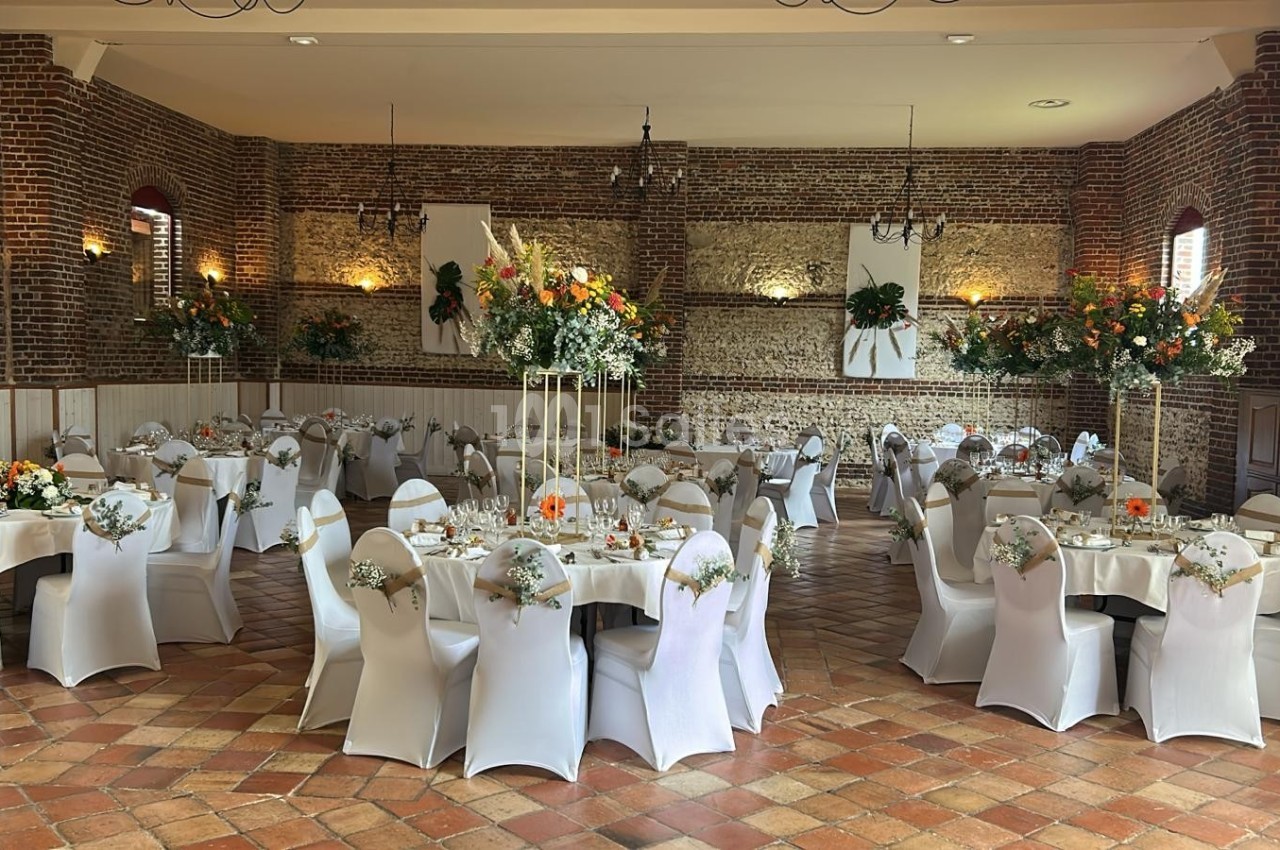 Salle de réception décorée avec des tables rondes, nappes blanches, chaises houssées et compositions florales élégantes.
