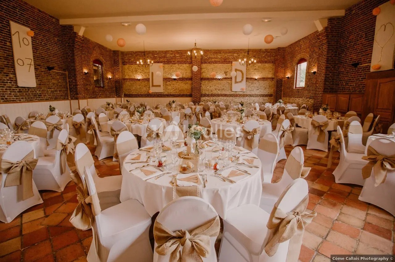 Salle de réception décorée pour un mariage, avec tables rondes dressées, chaises blanches et murs en briques apparentes.