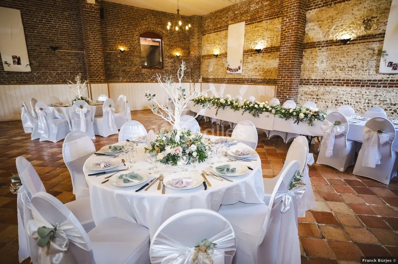 Salle de réception avec tables rondes décorées de nappes blanches, fleurs et vaisselle, dans un cadre en briques apparentes.