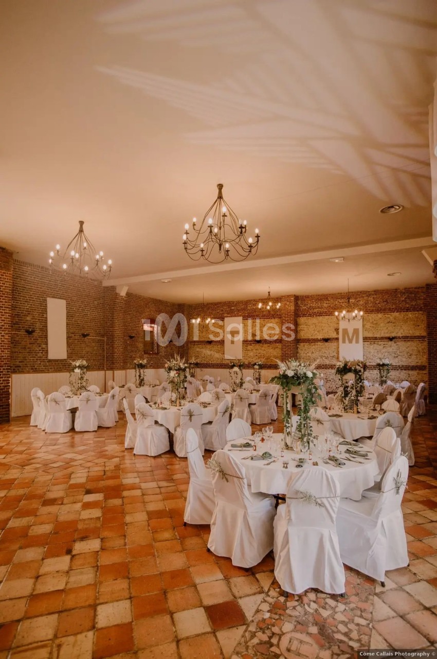 Salle de réception avec tables rondes décorées, chaises blanches et lustres suspendus, sur un sol en carreaux de terre cuite.