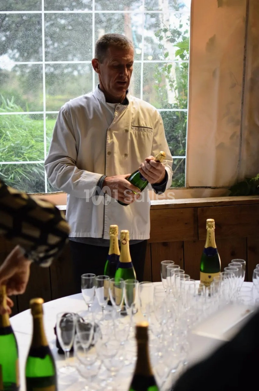 Un homme en veste de chef ouvre une bouteille de champagne devant une table garnie de verres et d'autres bouteilles.