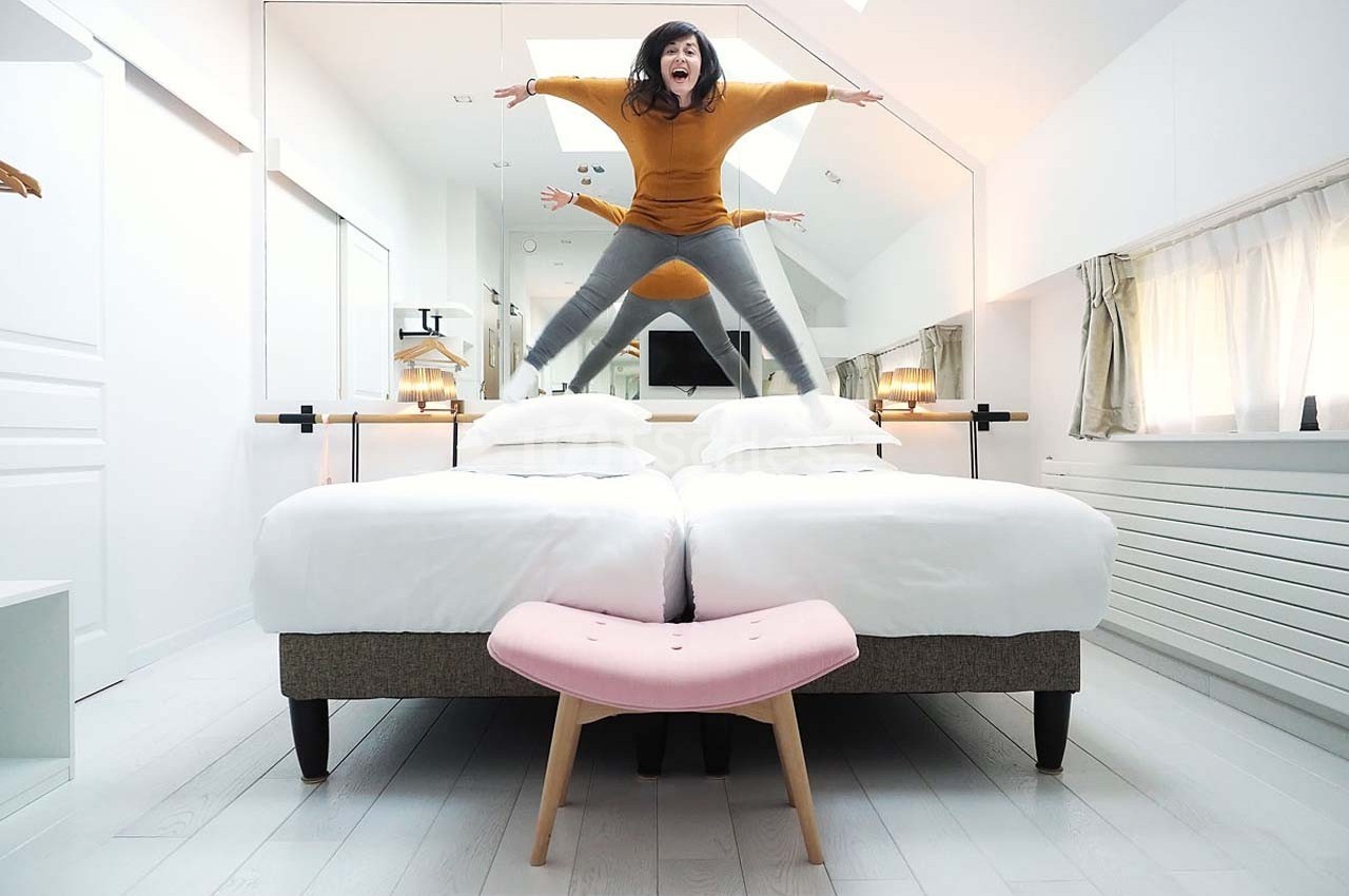 Une femme saute sur deux lits jumeaux dans une chambre lumineuse avec un miroir et un tabouret rose au premier plan.