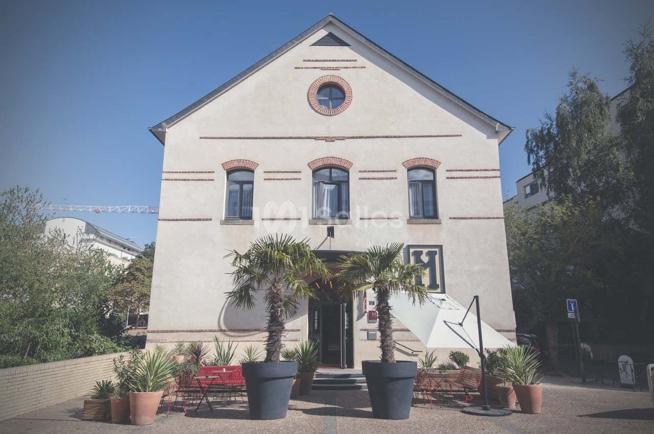 Façade d'un bâtiment clair à deux étages, entouré de plantes en pots et de bancs rouges sur une place pavée.