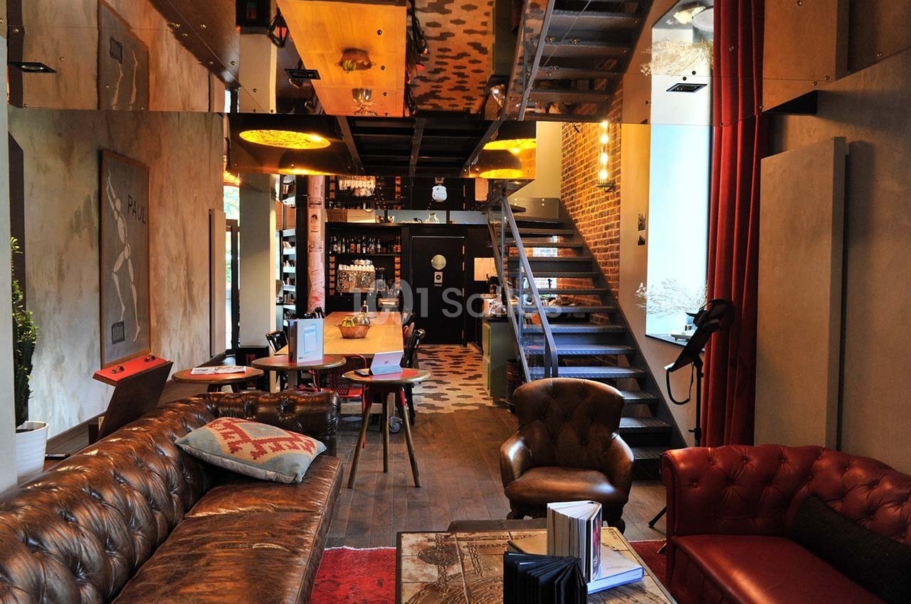 Salon chaleureux avec canapé en cuir, table basse, escalier métallique et bar en arrière-plan dans un décor moderne.