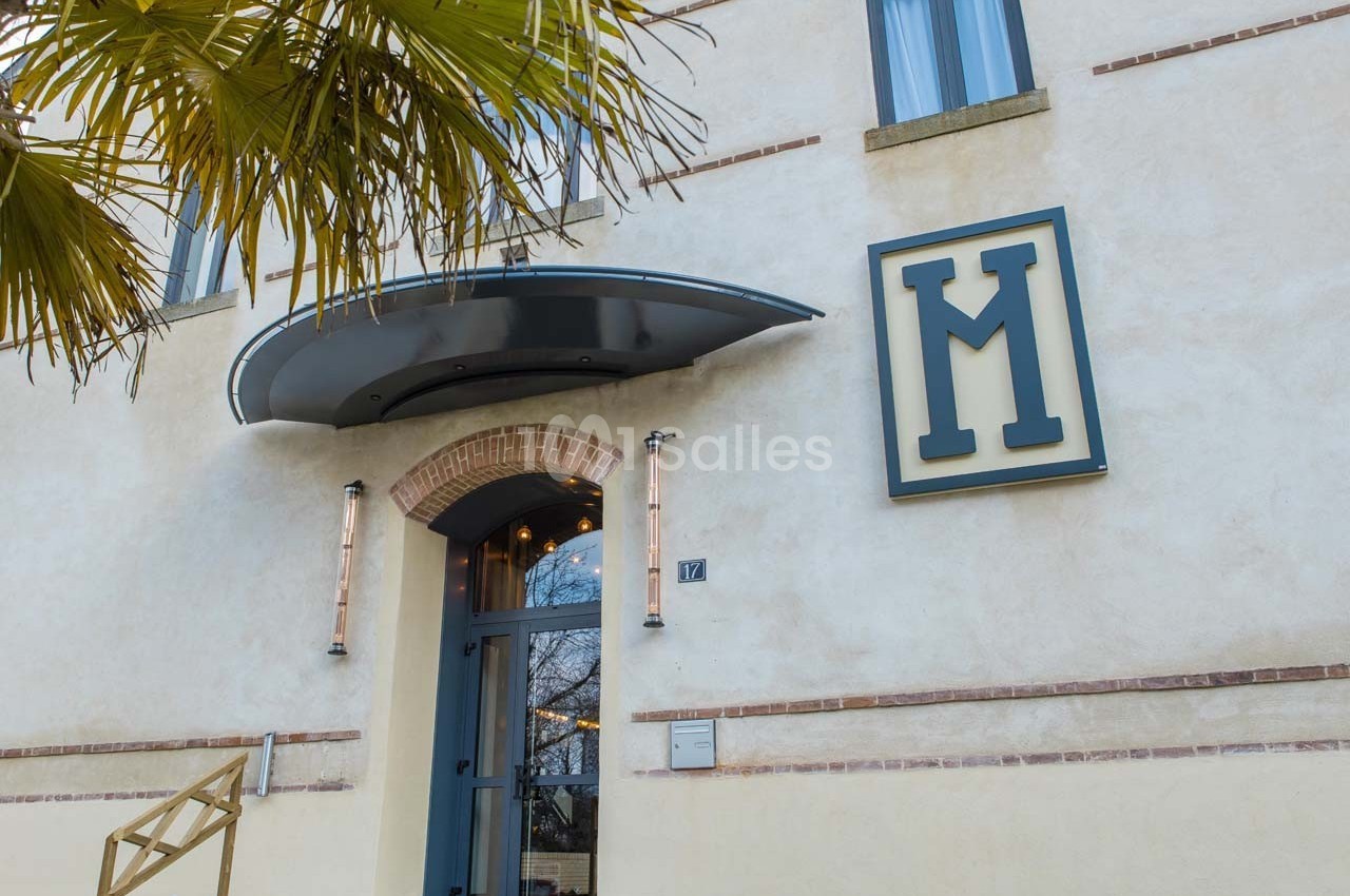 Façade d'un bâtiment beige avec une porte vitrée, un auvent métallique et un grand logo en forme de ’M’.