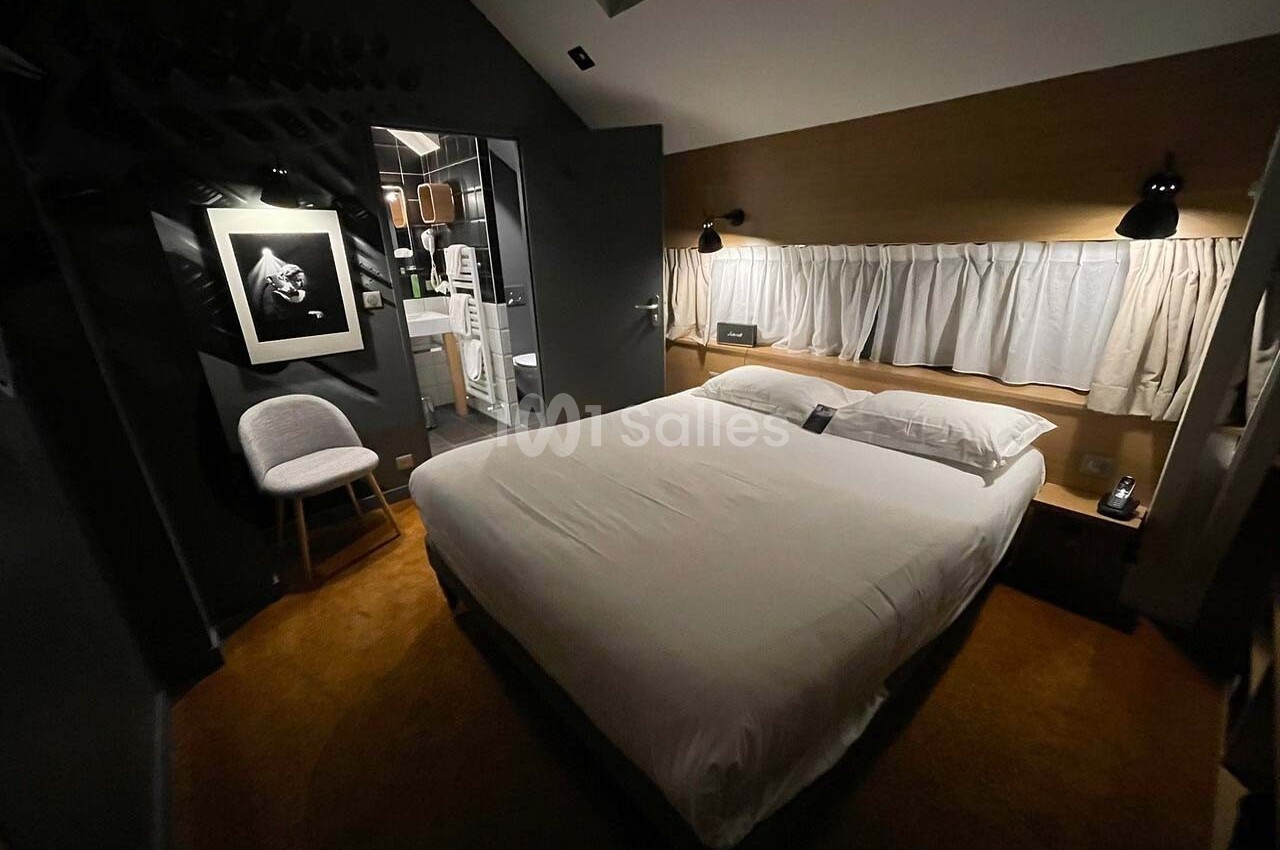 Chambre moderne avec lit double, éclairage tamisé, chaise, tableau au mur et salle de bain visible en arrière-plan.