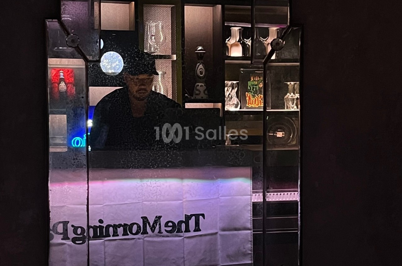 Un homme portant un chapeau est derrière un comptoir, reflété dans un miroir entouré d'étagères éclairées.