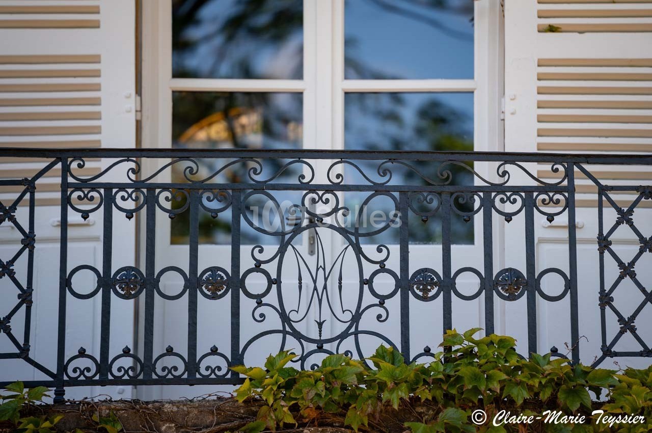Balustrade en fer forgé ornée de motifs circulaires et d'un monogramme, devant une fenêtre à volets blancs.