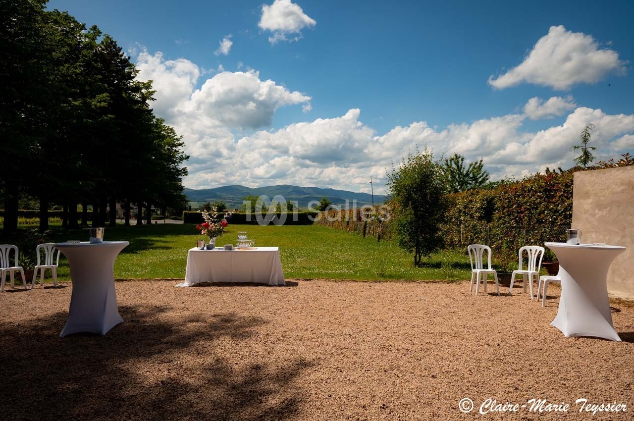 Jardin avec vue sur des collines, tables hautes blanches et table décorée de fleurs sur un sol gravillonné.