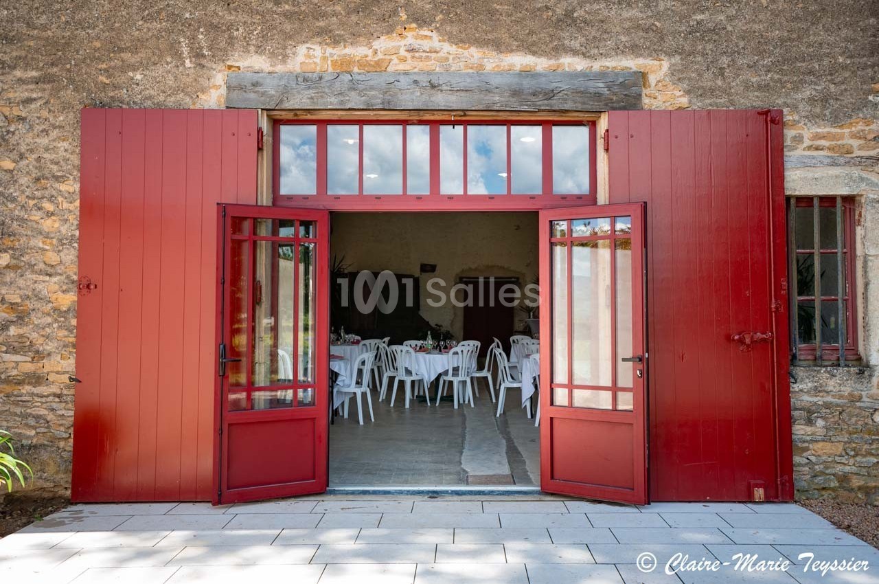 Portes rouges ouvertes sur une salle aménagée avec des chaises blanches, dans un bâtiment en pierre.