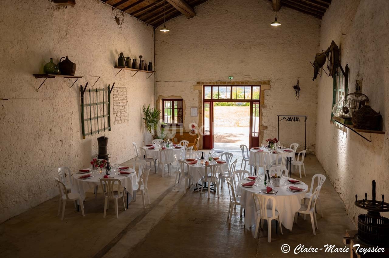 Salle rustique avec murs en pierre, tables rondes dressées avec nappes blanches et chaises, éclairée par des portes vitrées.
