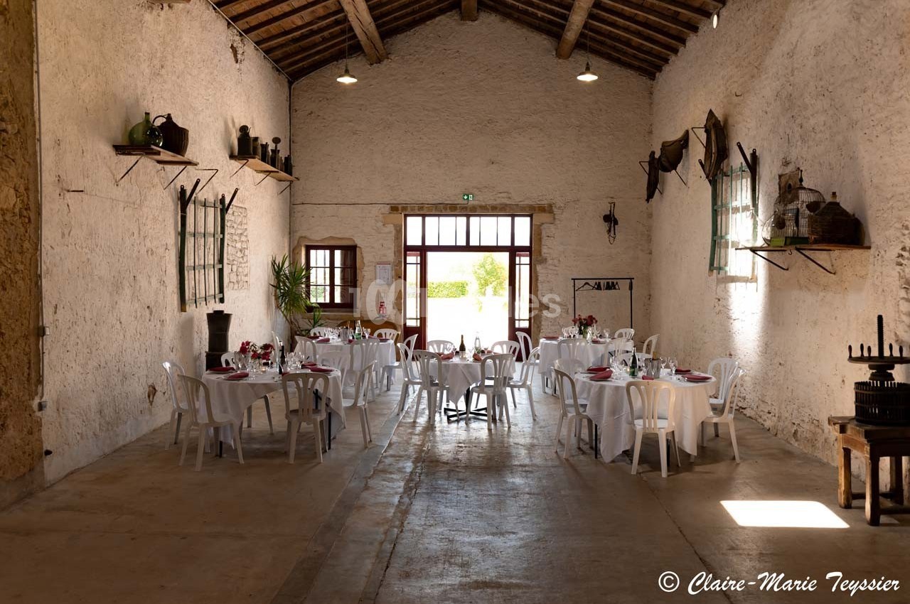 Salle de réception rustique avec tables rondes dressées, chaises blanches et lumière naturelle entrant par une grande porte.