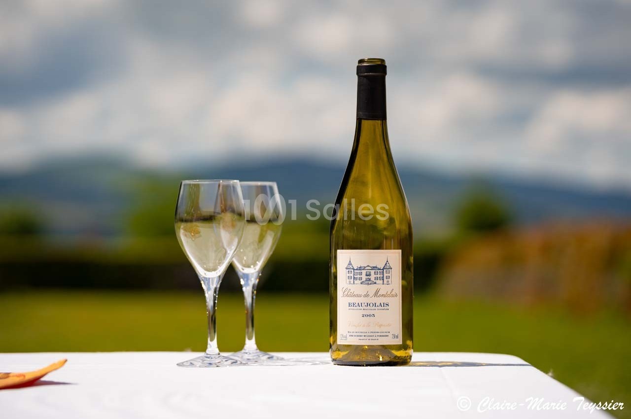 Bouteille de vin blanc et deux verres vides posés sur une table blanche en extérieur, avec un paysage flou en arrière-plan.