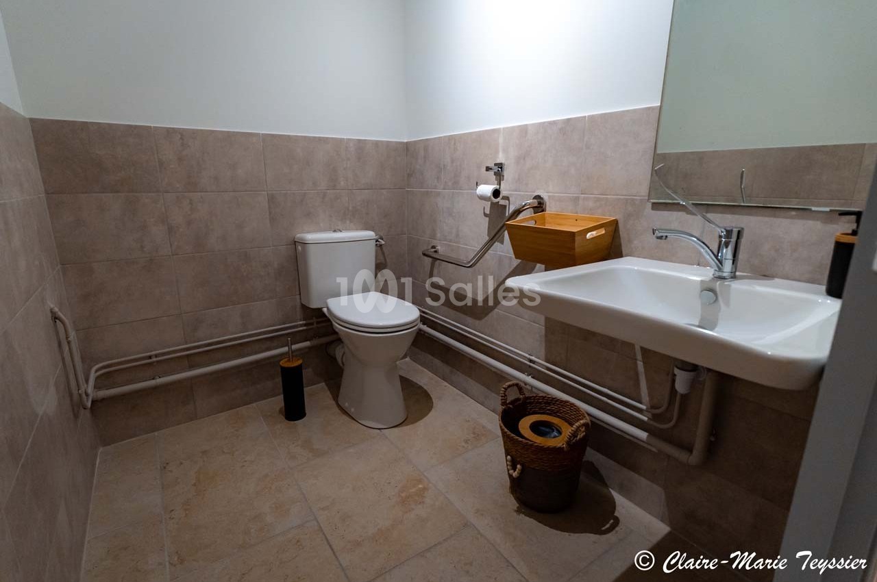 Toilettes accessibles avec lavabo, miroir, barre d'appui et carrelage beige au mur et au sol.