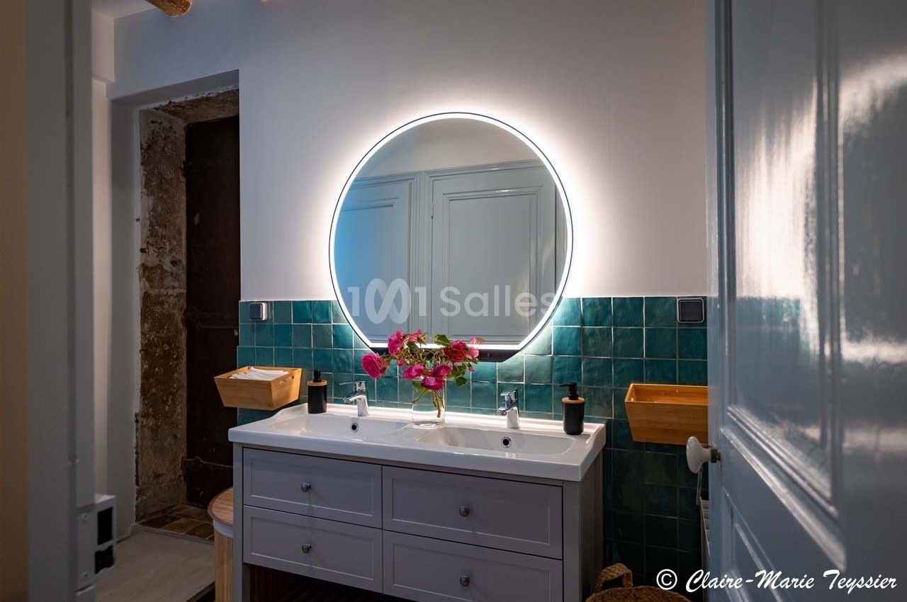 Salle de bain avec un miroir rond éclairé, un plan vasque double blanc et des carreaux muraux verts.