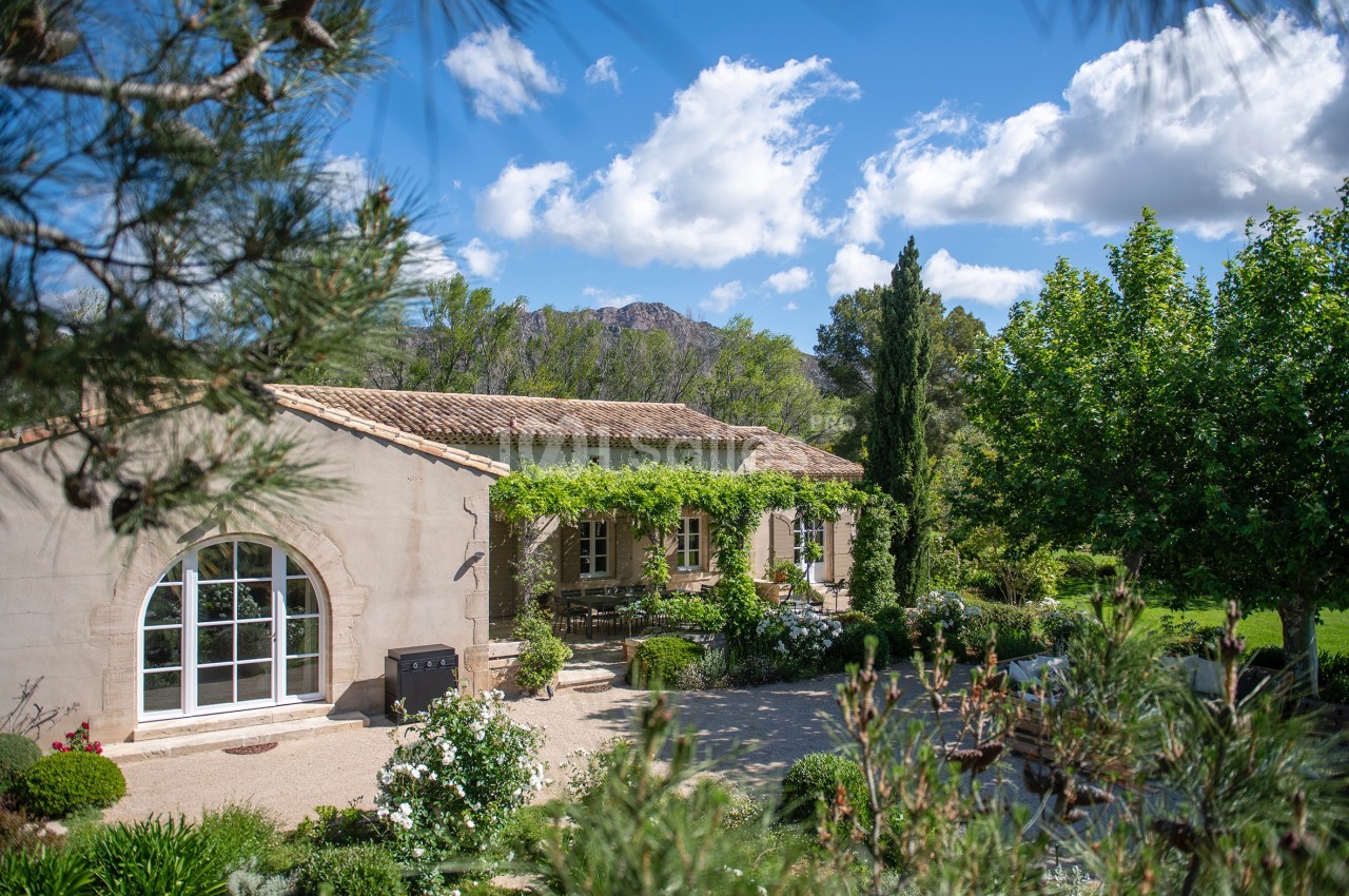 Maison en pierre entourée de verdure avec terrasse ombragée, jardin fleuri et montagnes en arrière-plan.