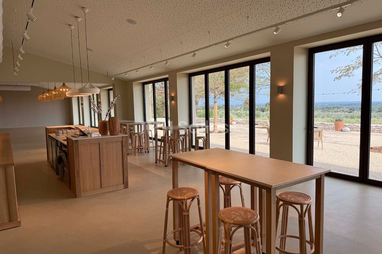 Salle lumineuse avec tables en bois, tabourets et grandes baies vitrées donnant sur un paysage extérieur.