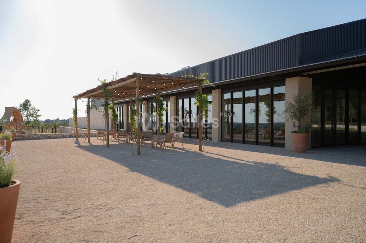 Terrasse ombragée avec pergola en bois, tables et chaises, devant un bâtiment moderne avec grandes baies vitrées.
