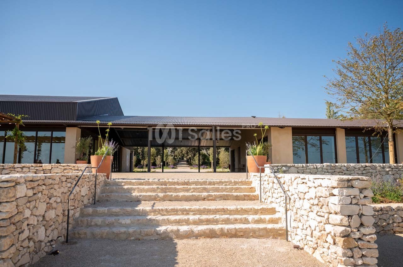 Entrée d'un bâtiment moderne avec toit en métal, entourée de murs en pierre et de plantes en pots sous un ciel dégagé.
