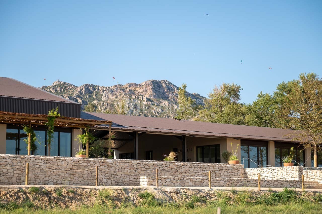 Bâtiment moderne avec terrasse en bois, entouré de végétation, au pied d'une montagne sous un ciel dégagé.