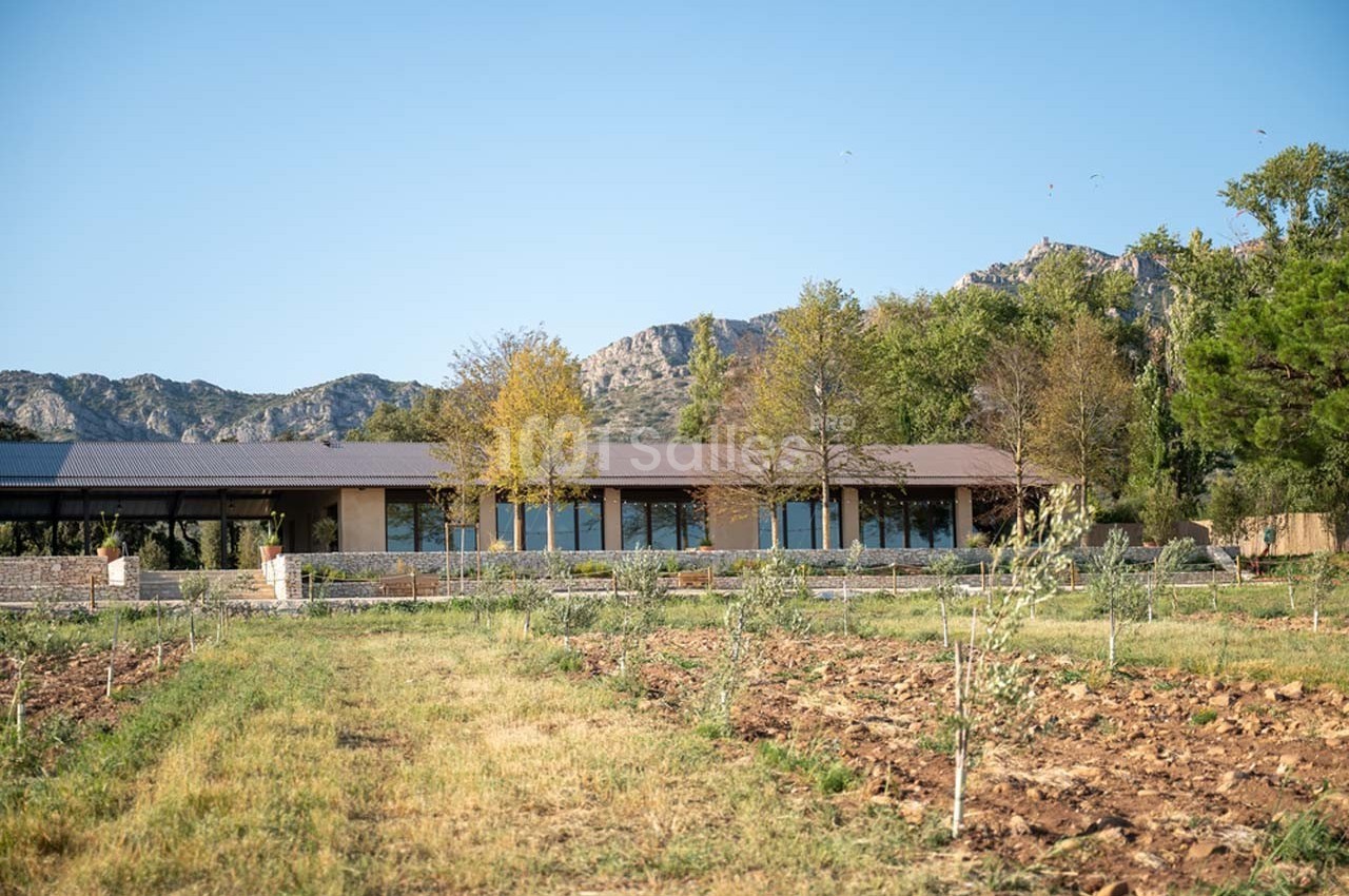 Bâtiment moderne entouré de champs cultivés, avec des montagnes et des arbres en arrière-plan sous un ciel dégagé.