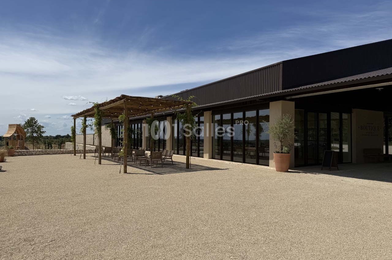 Bâtiment moderne avec terrasse ombragée par une pergola, entouré d'un espace extérieur en gravier sous un ciel dégagé.