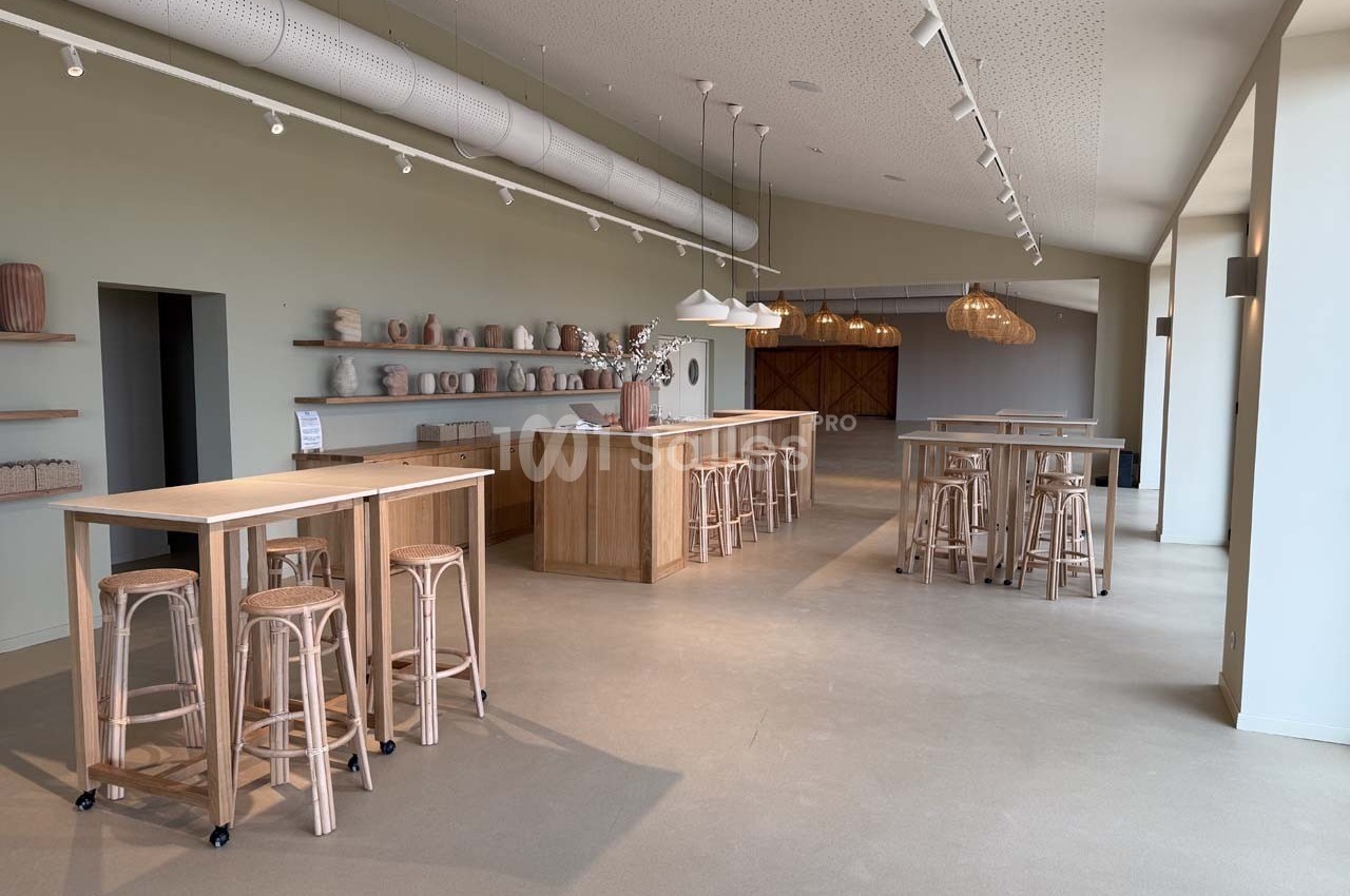 Salle lumineuse avec tables hautes en bois, tabourets, étagères décorées de céramiques et suspensions en osier.