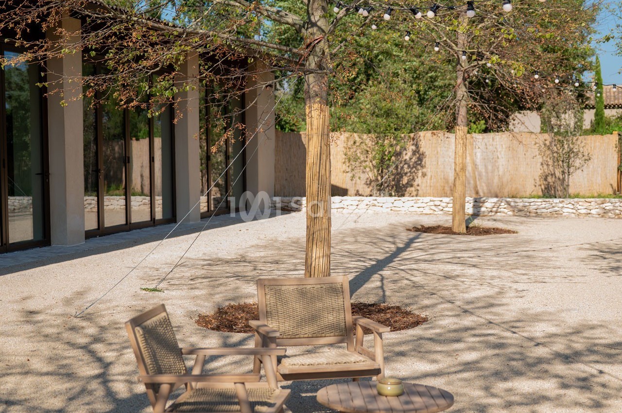 Mobilier de jardin en bois sous des arbres avec guirlandes lumineuses, dans une cour ensoleillée et gravillonnée.