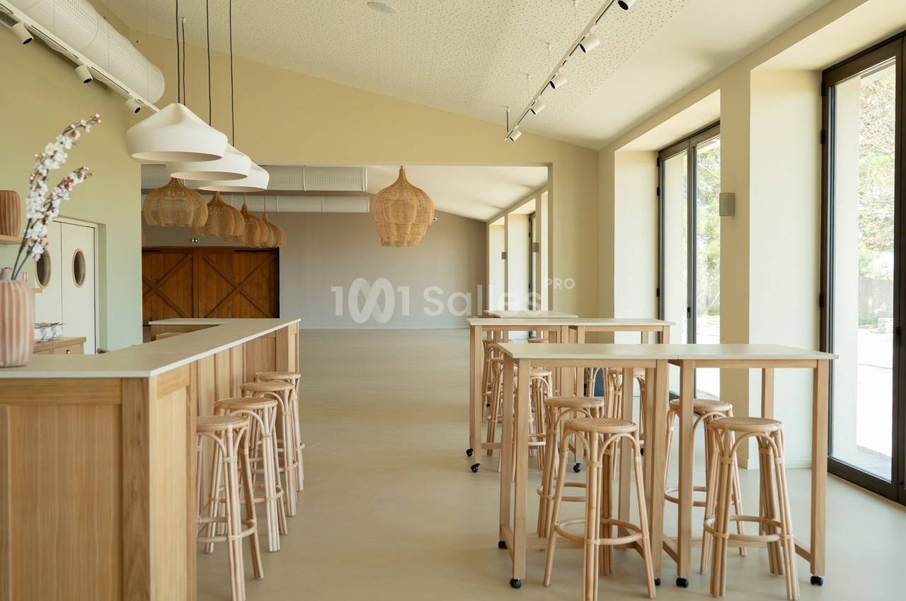 Salle lumineuse avec tables hautes en bois, tabourets assortis et suspensions en osier, grandes baies vitrées sur la droite.