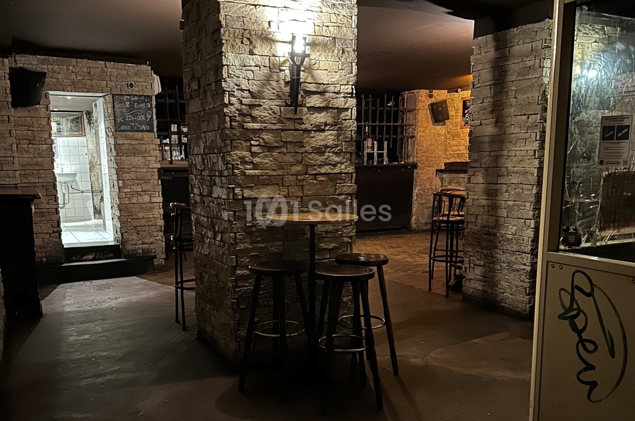 Intérieur d'un bar sombre avec des murs en pierre, des tabourets autour d'une table et un éclairage tamisé.