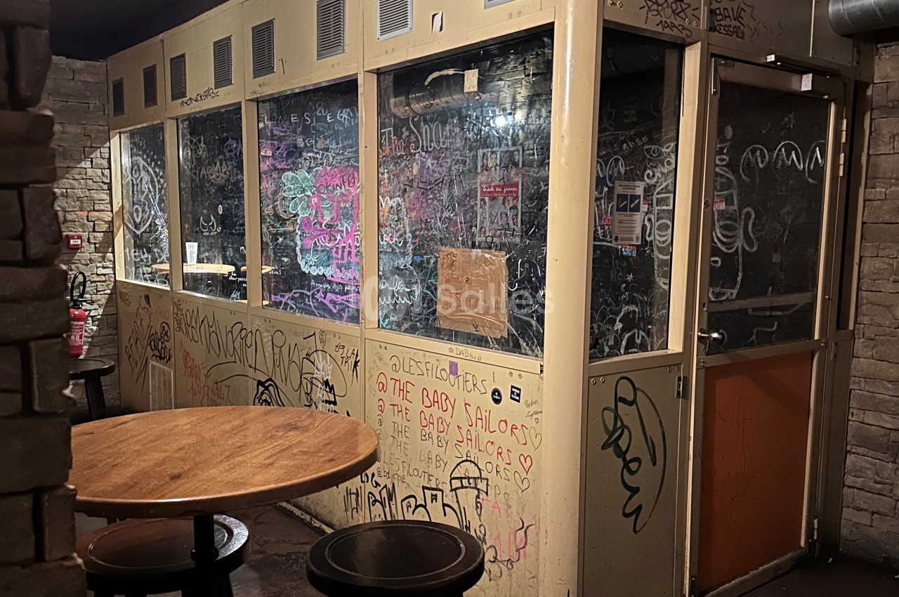 Cabine vitrée dans un espace intérieur, recouverte de graffitis et tags, avec une table ronde au premier plan.