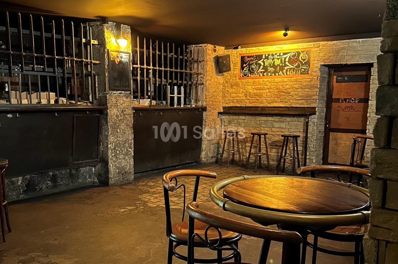 Salle de bar sombre avec tables en bois, tabourets hauts, mur en briques et tableau noir décoratif.
