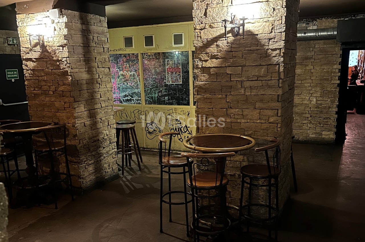 Salle avec murs en pierre, tables hautes en bois et graffitis sur une vitre au fond, éclairée par des appliques murales.
