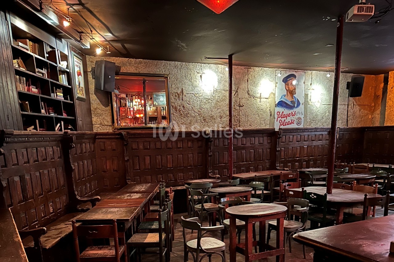Salle de bar rustique avec tables en bois, étagères de livres, éclairage tamisé et affiche murale décorative.